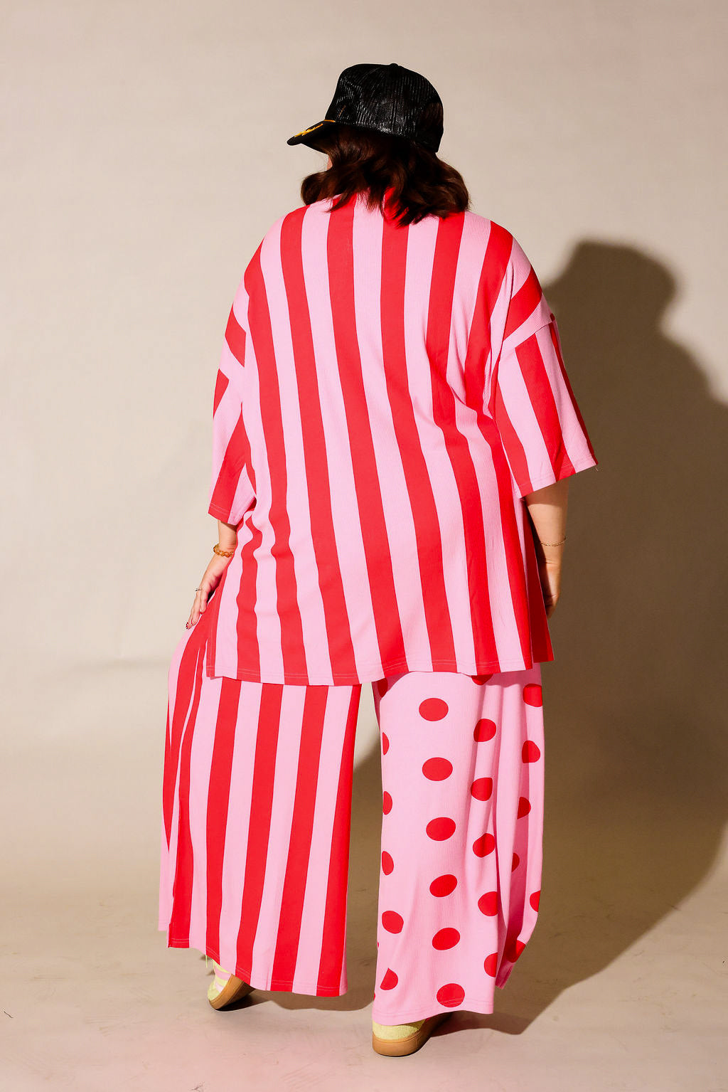 Model wearing a red and white striped set with a black trucker at on a beige background, back view
