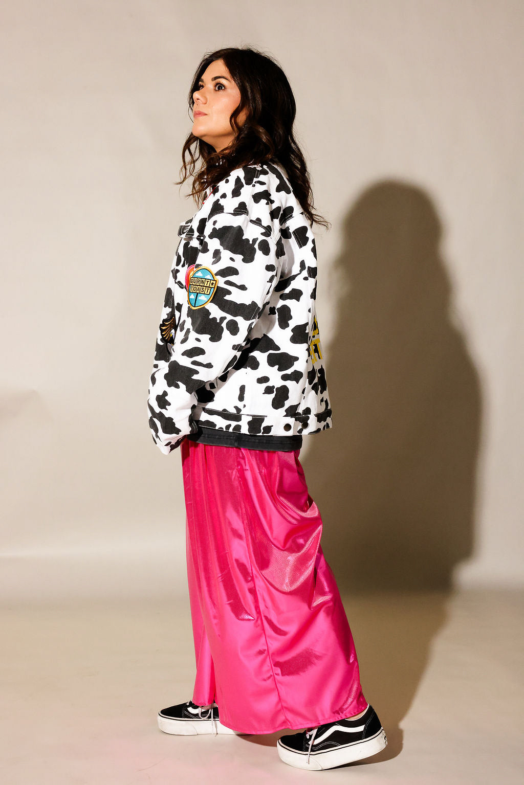 A woman stands in profile wearing a white and black cow-print jacket with colorful patches, paired with shiny hot pink wide-leg pants and black sneakers, against a beige studio background with a cast shadow. side view