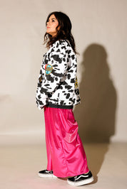 A woman stands in profile wearing a white and black cow-print jacket with colorful patches, paired with shiny hot pink wide-leg pants and black sneakers, against a beige studio background with a cast shadow. side view