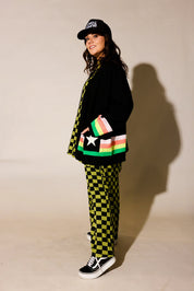 Person wearing a black jacket, checkered pants, and colorful bag against a beige background, side view