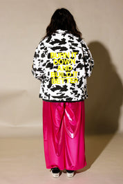 A person shown from the back wearing a black and white cow-print jacket with bold yellow text reading 'BURN IT DOWN AND BUILD IT BETTER,' paired with shiny hot pink wide-leg pants and black sneakers, against a plain beige studio backdrop. back view