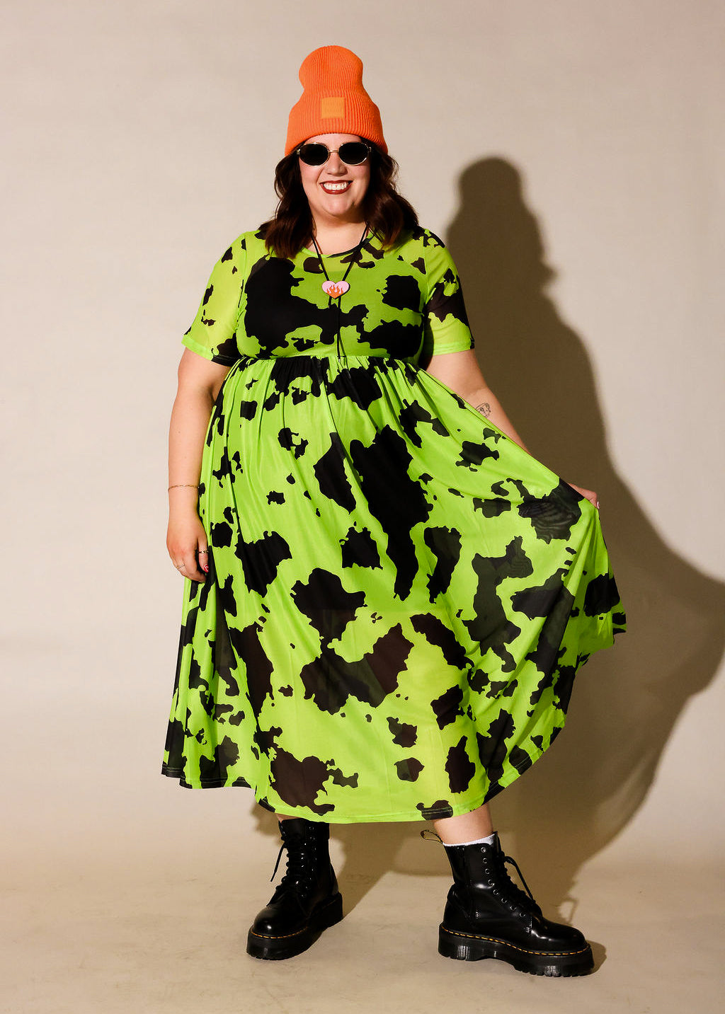 Person wearing a bright green dress with black patterns, black boots, and an orange beanie against a beige background, frontal view