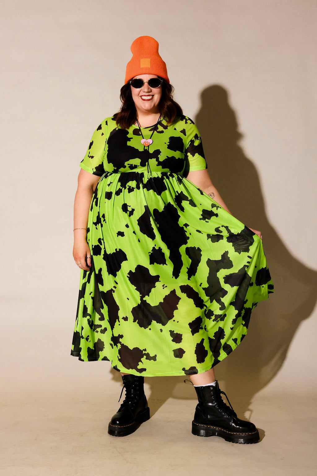 Person wearing a bright green dress with black patterns, black boots, and an orange beanie against a beige background, frontal view