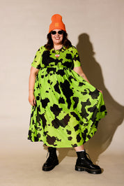 Person wearing a bright green dress with black patterns, black boots, and an orange beanie against a beige background, frontal view