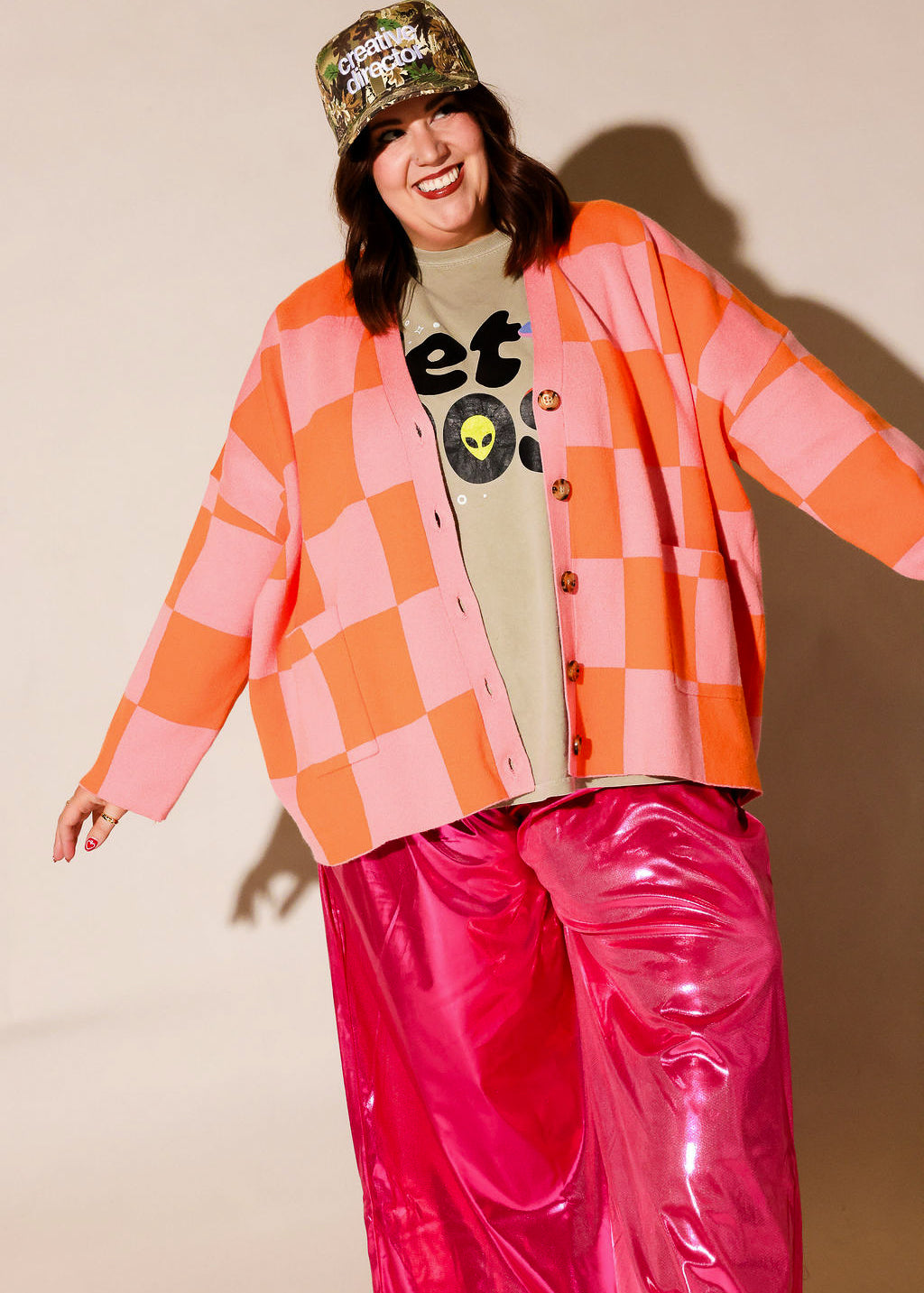 A person modeling a bright outfit against a plain beige studio background, wearing a pink and orange checkered cardigan, a beige graphic T-shirt, shiny hot pink pants, and a camouflage cap with text, frontal view