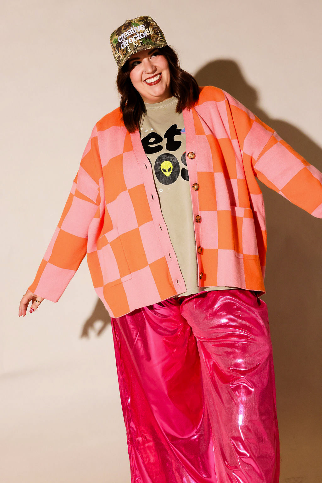 A person modeling a bright outfit against a plain beige studio background, wearing a pink and orange checkered cardigan, a beige graphic T-shirt, shiny hot pink pants, and a camouflage cap with text, frontal view