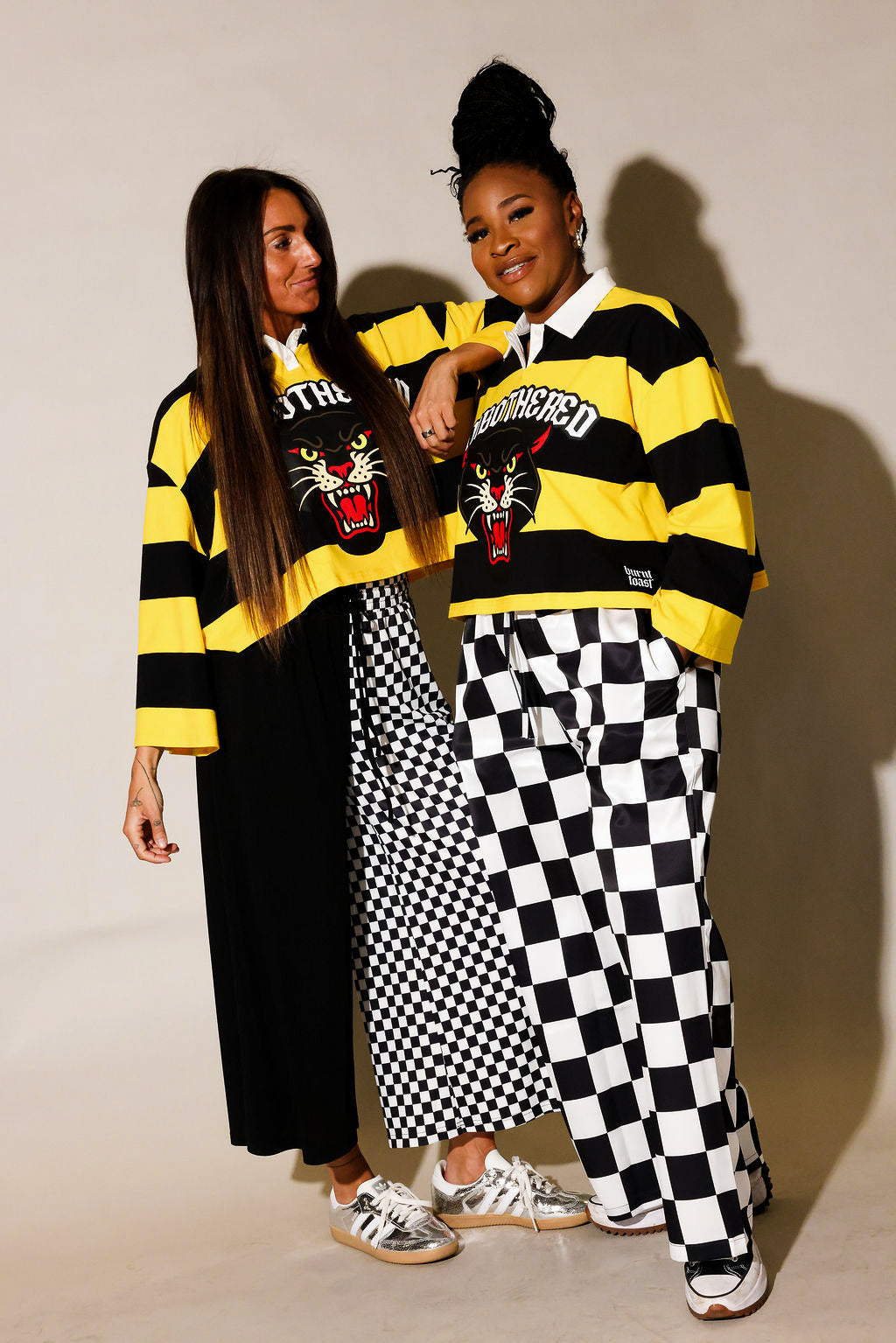 Two models wearing yellow and black polos and black and white pants, standing against a beige background, frontal view