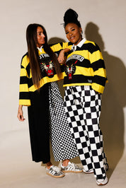 Two models wearing yellow and black polos and black and white pants, standing against a beige background, frontal view