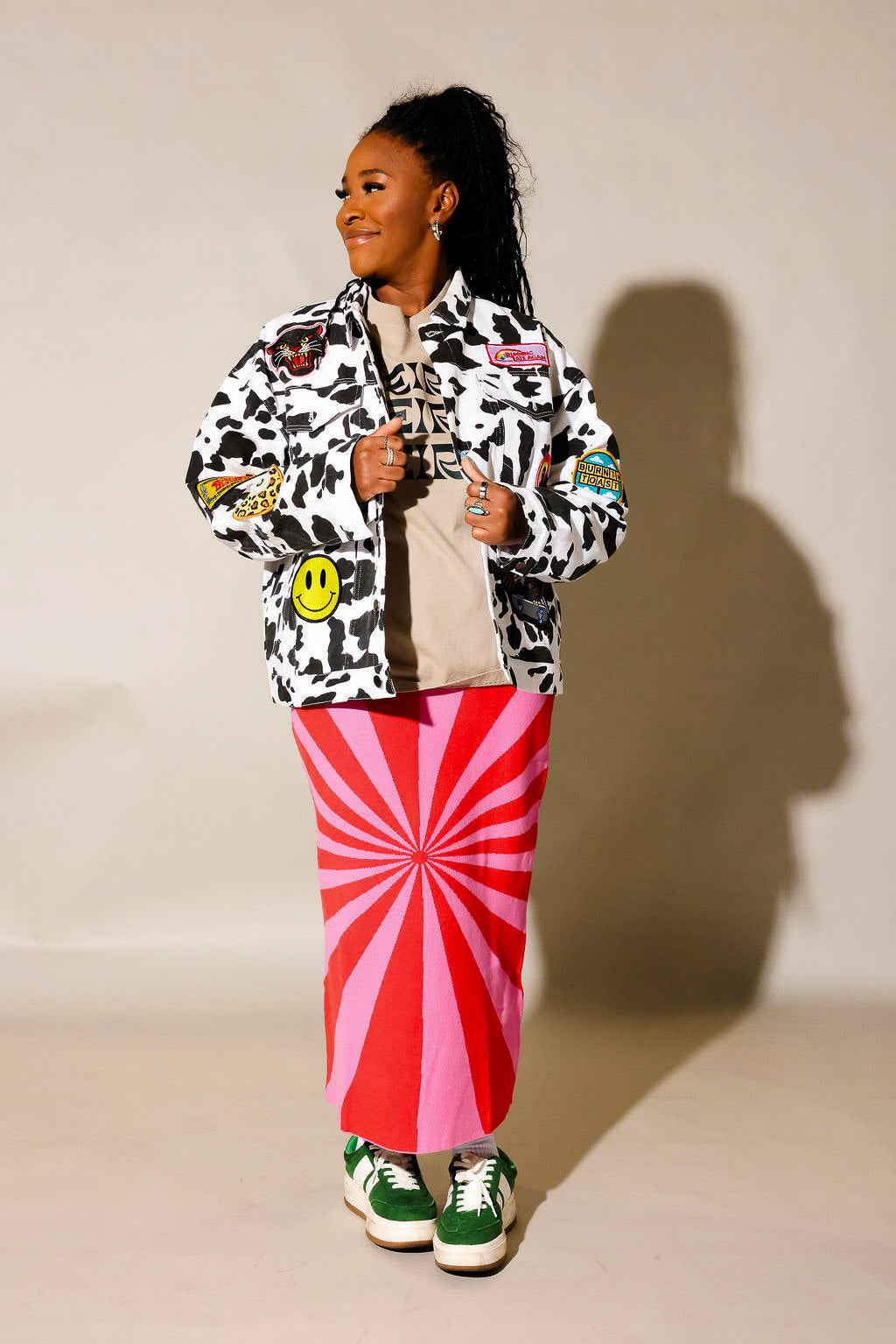 Model wearing a red and pink spiral patterned knit skirt, and cow pattern denim jacket, against a beige wall, frontal view