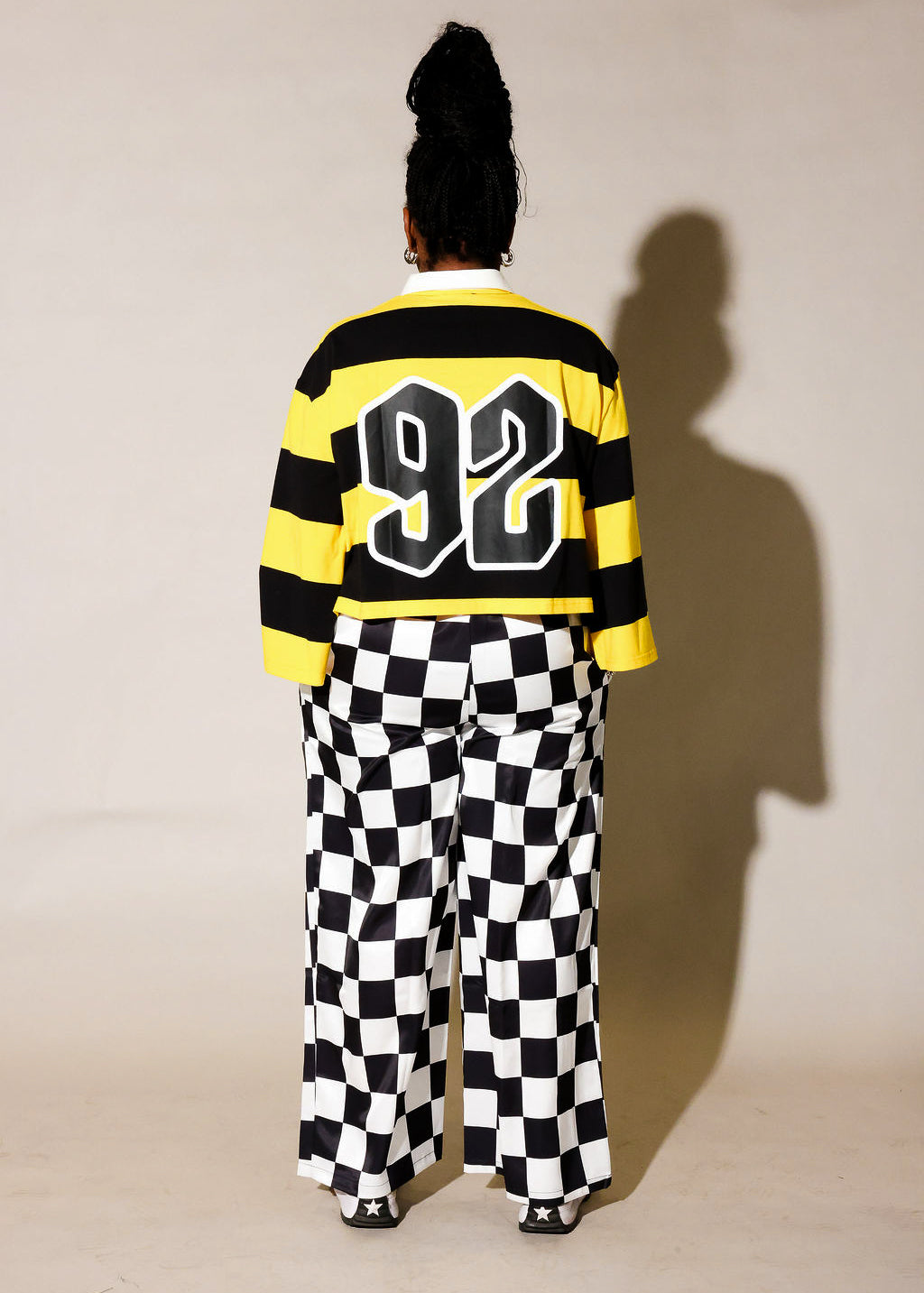 Model wearing a black and yellow striped polo with 92 on the back, and black and white checkered pants, back view