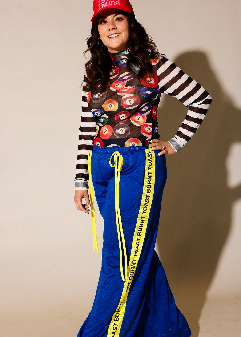 A model posing in a streetwear outfit featuring bright blue pants with a yellow 'TOAST BURNT TOAST' stripe, a black and white striped long-sleeve shirt, a colorful patterned crop top, and a red baseball cap, frontal view