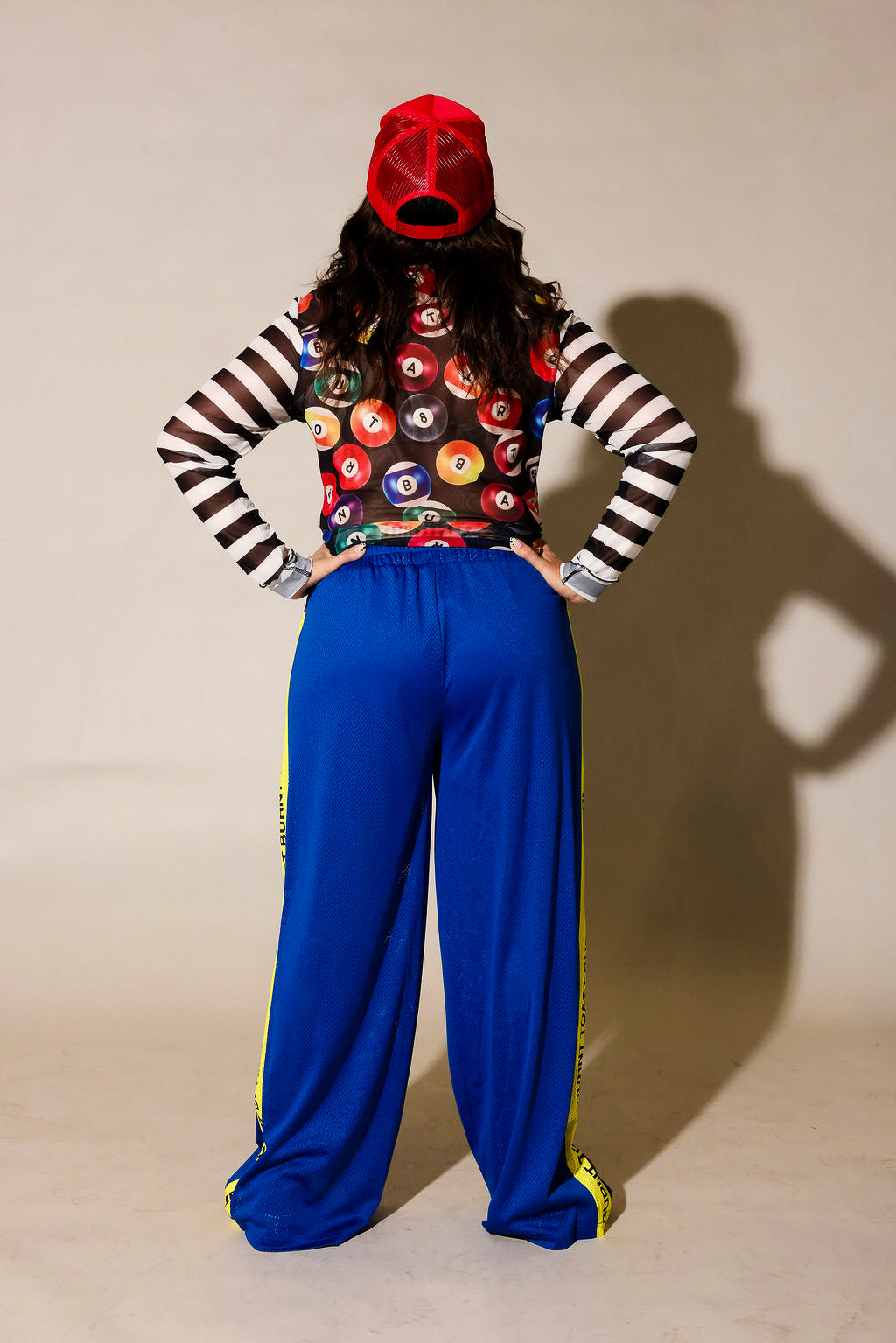 A model posing in a streetwear outfit featuring bright blue pants with a yellow 'TOAST BURNT TOAST' stripe, a black and white striped long-sleeve shirt, a colorful patterned crop top, and a red baseball cap, back view