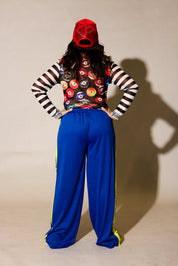 A model posing in a streetwear outfit featuring bright blue pants with a yellow 'TOAST BURNT TOAST' stripe, a black and white striped long-sleeve shirt, a colorful patterned crop top, and a red baseball cap, back view