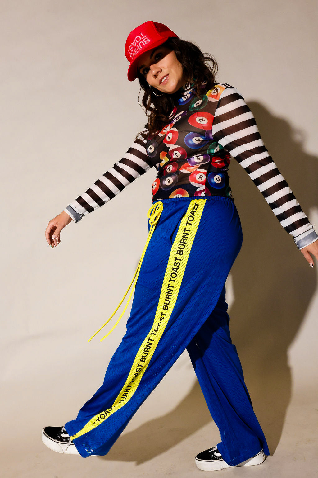 A model posing in a streetwear outfit featuring bright blue pants with a yellow 'TOAST BURNT TOAST' stripe, a black and white striped long-sleeve shirt, a colorful patterned crop top, and a red baseball cap, side view