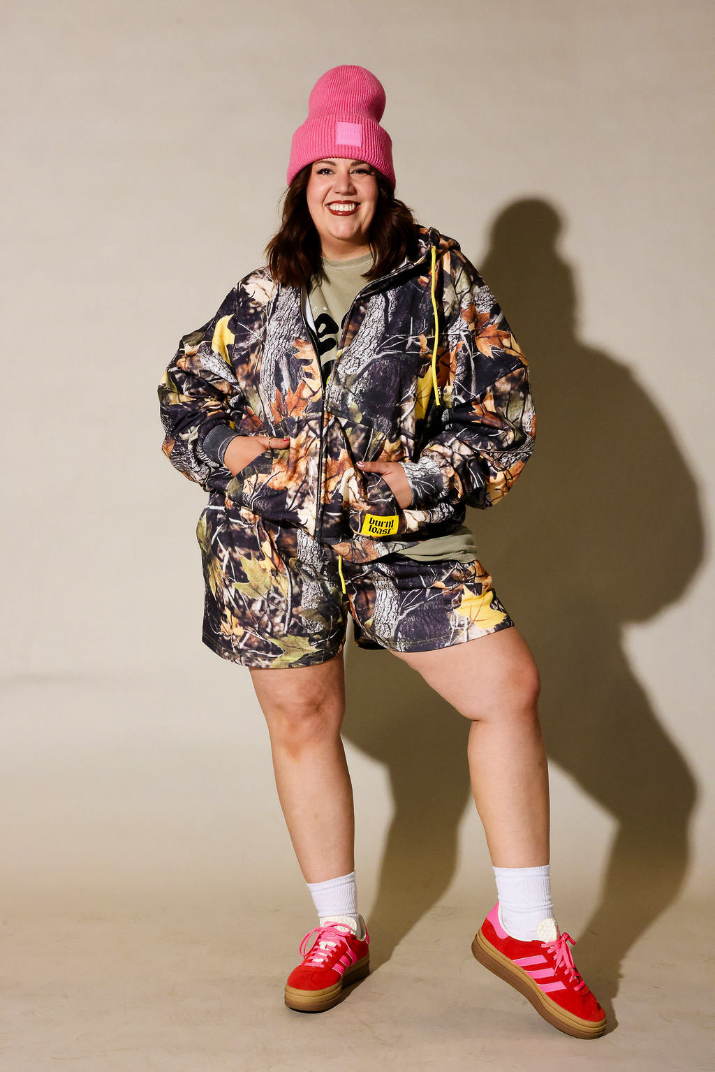 A woman modeling a matching camouflage print hooded jacket and shorts set, styled with a bright pink beanie, a green t-shirt, white socks, and red sneakers against a beige studio background, frontal view