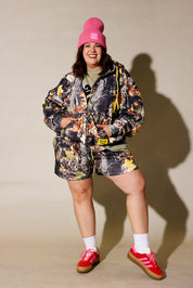 A woman modeling a matching camouflage print hooded jacket and shorts set, styled with a bright pink beanie, a green t-shirt, white socks, and red sneakers against a beige studio background, frontal view