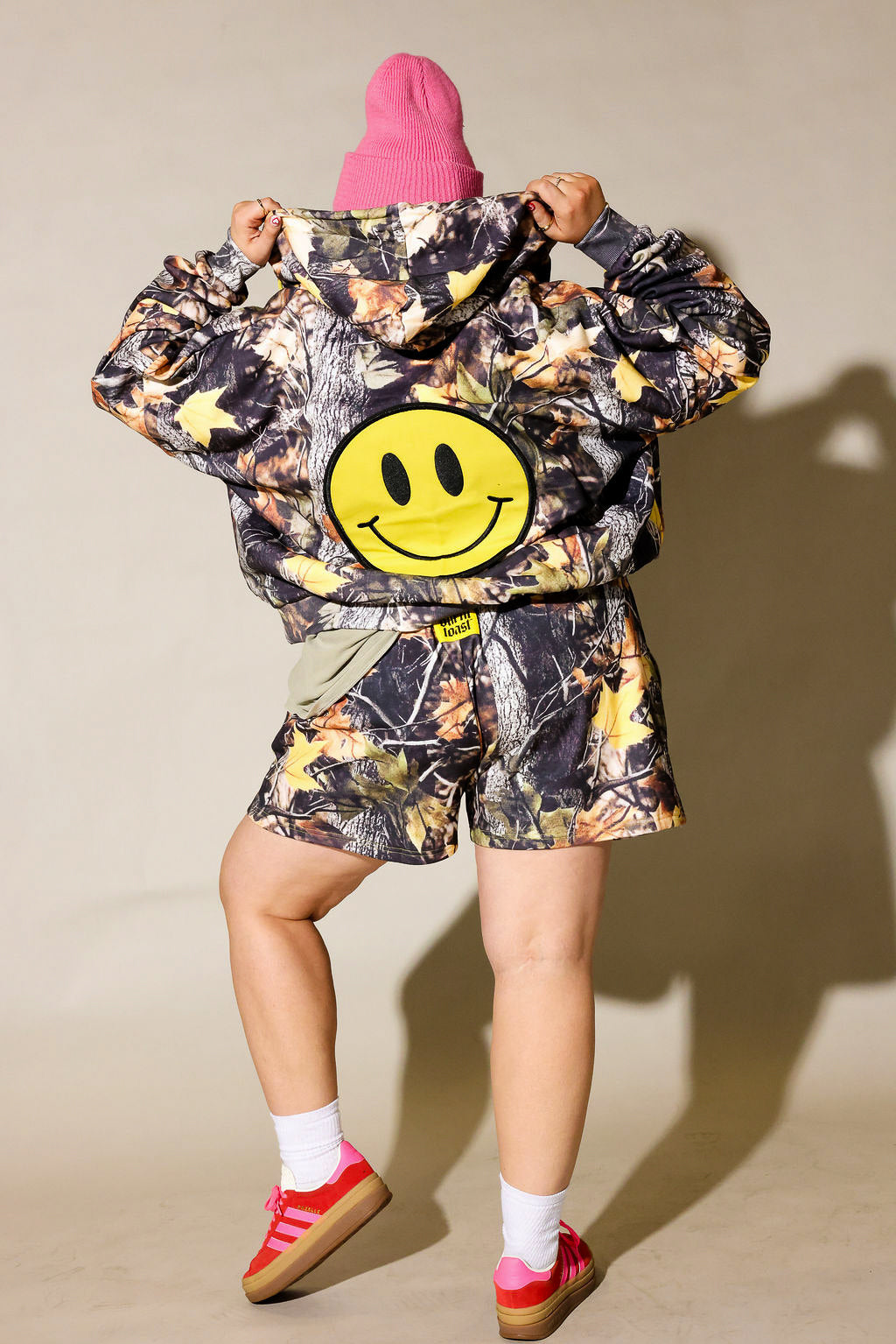 A woman modeling a matching camouflage print hooded jacket and shorts set, styled with a bright pink beanie, a green t-shirt, white socks, and red sneakers against a beige studio background, back view