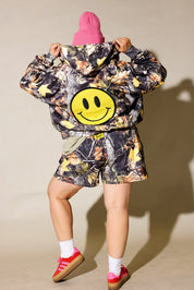 A woman modeling a matching camouflage print hooded jacket and shorts set, styled with a bright pink beanie, a green t-shirt, white socks, and red sneakers against a beige studio background, back view