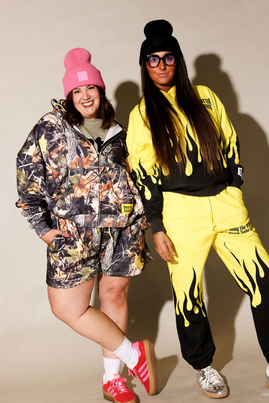 Two models posing in matching streetwear tracksuits. The model on the left wears a camouflage-patterned hooded jacket and shorts with a pink beanie and red sneakers, frontal view