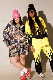 Two models posing in matching streetwear tracksuits. The model on the left wears a camouflage-patterned hooded jacket and shorts with a pink beanie and red sneakers, frontal view