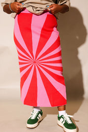 A person wearing a long midi skirt with a bold red and pink sunburst pattern, styled with a beige top, white socks, and green sneakers against a plain beige background, close up frontal view