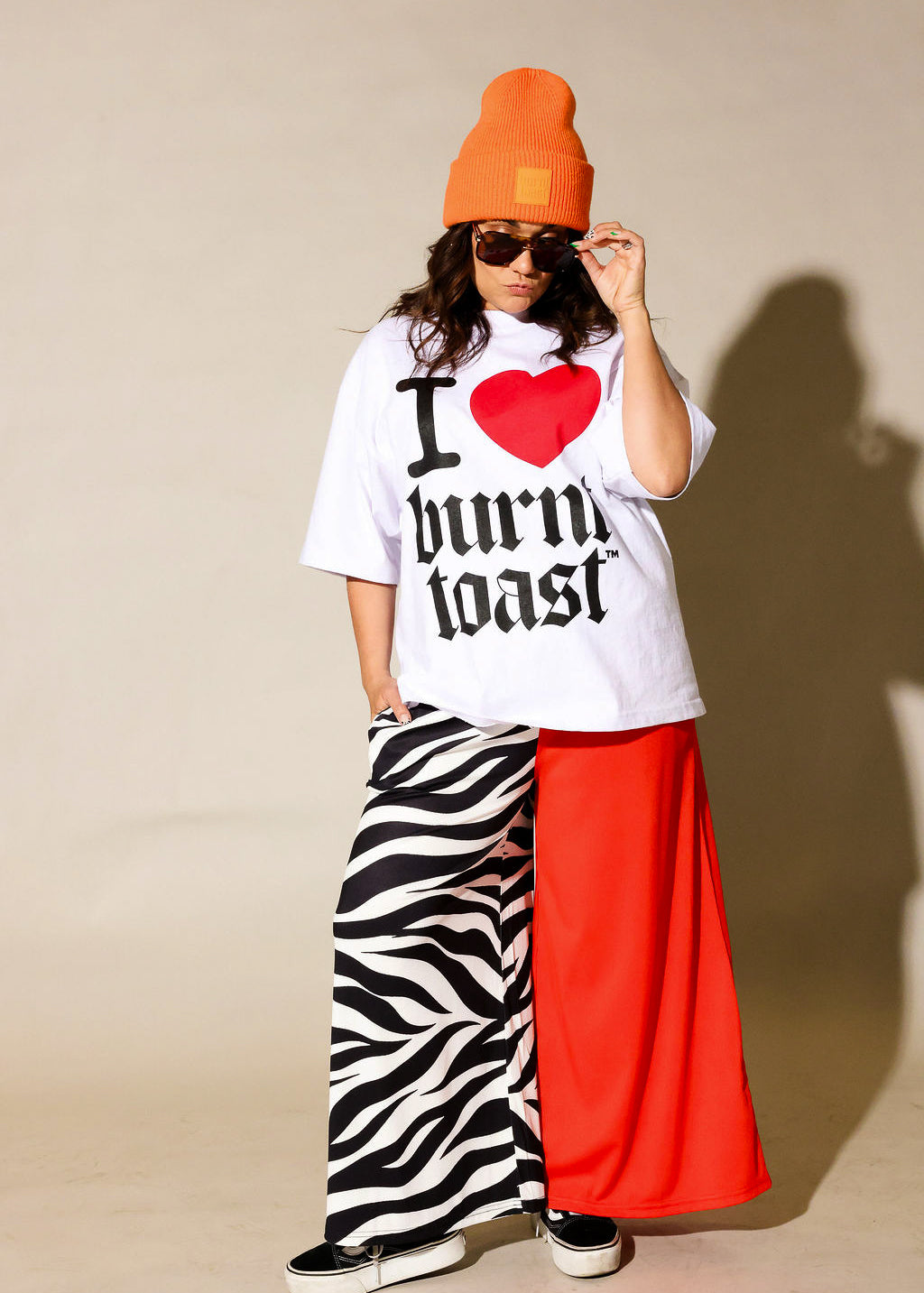 Person wearing a white t-shirt with text, orange beanie, zebra print and red pants against a beige background, frontal view