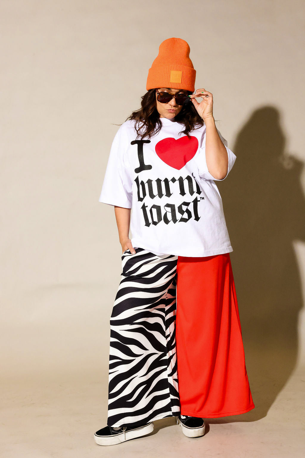 Person wearing a white t-shirt with text, orange beanie, zebra print and red pants against a beige background, frontal view