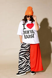 Person wearing a white t-shirt with text, orange beanie, zebra print and red pants against a beige background, frontal view