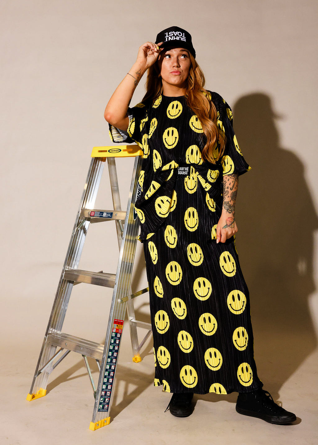 Model wearing a black short sleeve and long skirt plisse set with yellow smiley face patterns, black trucker hat with white upside down 'burnt toast' logo, black tennis shoes, standing next to a ladder, frontal view