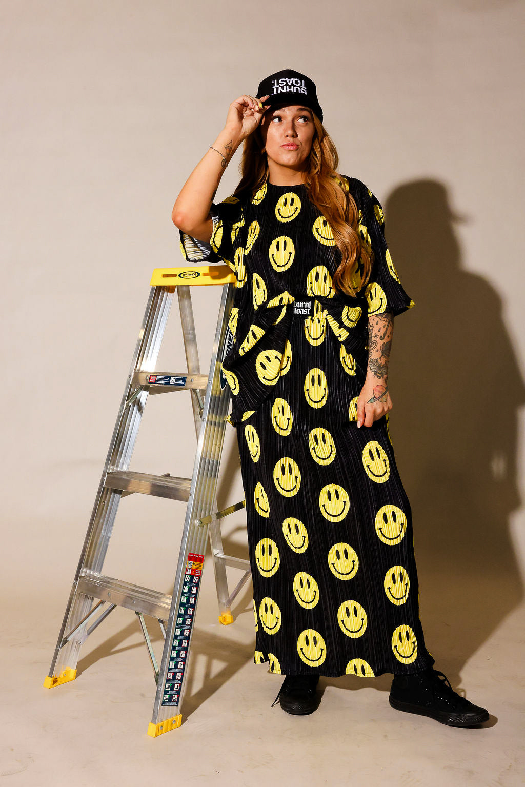 Model wearing a black short sleeve and long skirt plisse set with yellow smiley face patterns, black trucker hat with white upside down 'burnt toast' logo, black tennis shoes, standing next to a ladder, frontal view