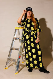 Model wearing a black short sleeve and long skirt plisse set with yellow smiley face patterns, black trucker hat with white upside down 'burnt toast' logo, black tennis shoes, standing next to a ladder, frontal view