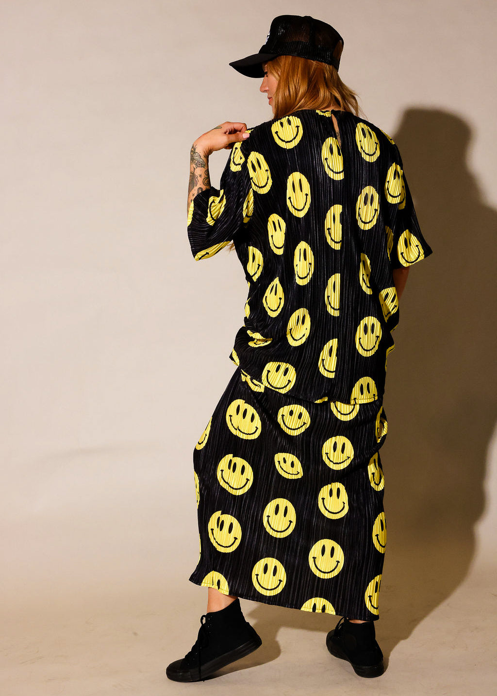 Model wearing a black short sleeve and long skirt plisse set with yellow smiley face patterns, black trucker hat with white upside down 'burnt toast' logo, black tennis shoes, standing, back view