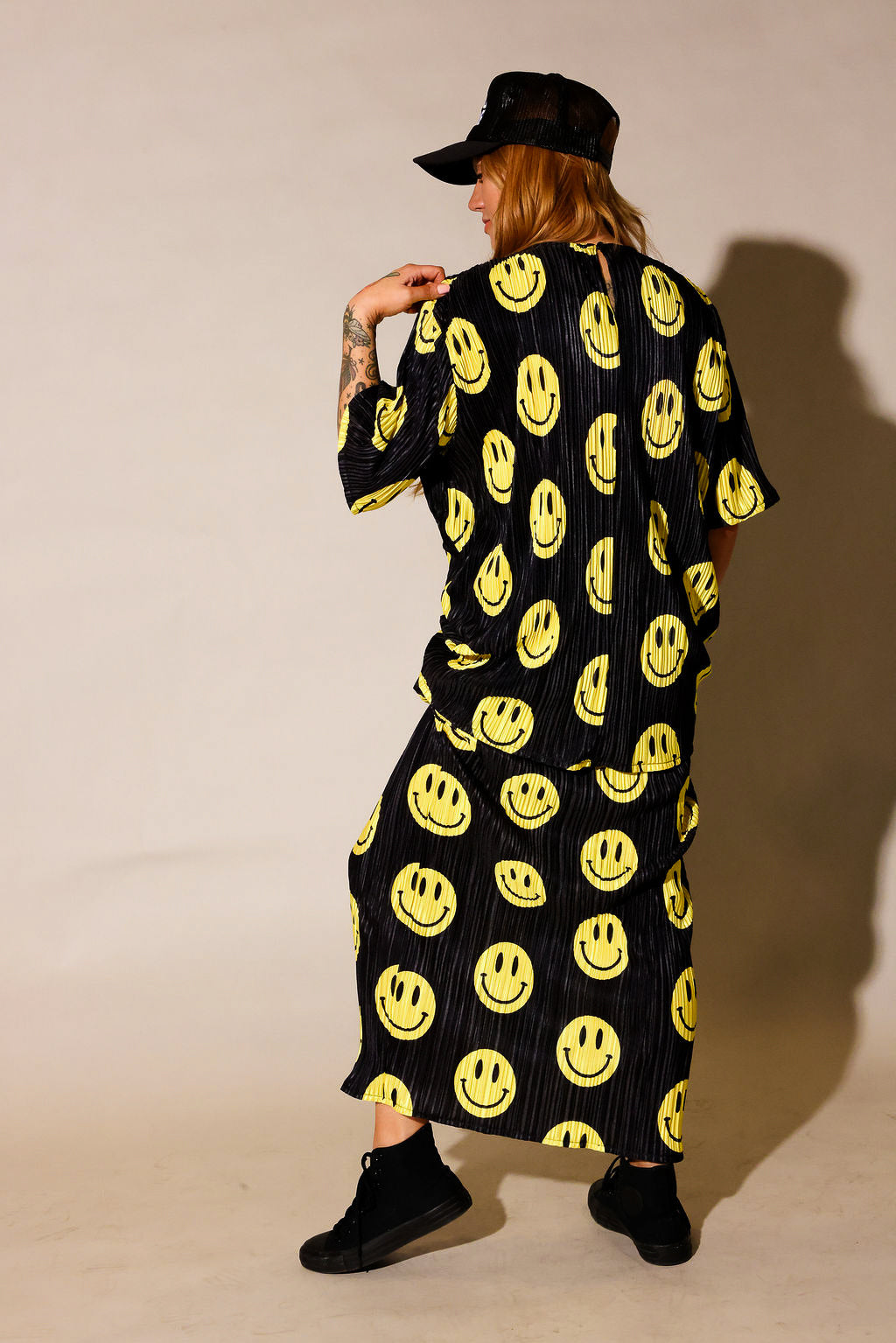 Model wearing a black short sleeve and long skirt plisse set with yellow smiley face patterns, black trucker hat with white upside down 'burnt toast' logo, black tennis shoes, standing, back view