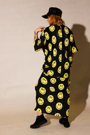 Model wearing a black short sleeve and long skirt plisse set with yellow smiley face patterns, black trucker hat with white upside down 'burnt toast' logo, black tennis shoes, standing, back view