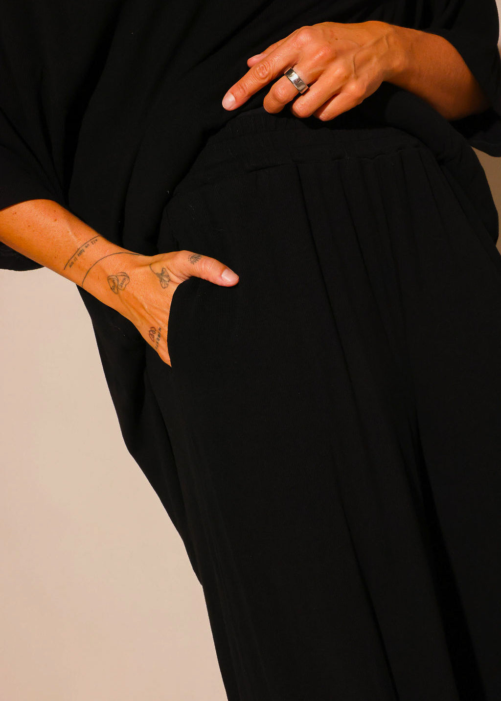 Close up of a model wearing a black ribbed two piece set, showing a hand in the pocket of the high waisted pants and the relaxed fit of the outfit