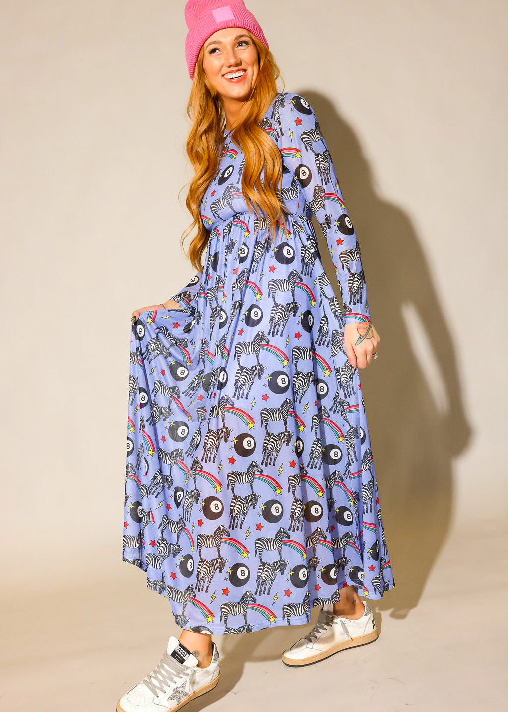 Model wearing a pink beanie, long purple mesh dress with a zebra, rainbow and 8 ball design print, and white sneakers with a silver star, against a beige background, full frontal view