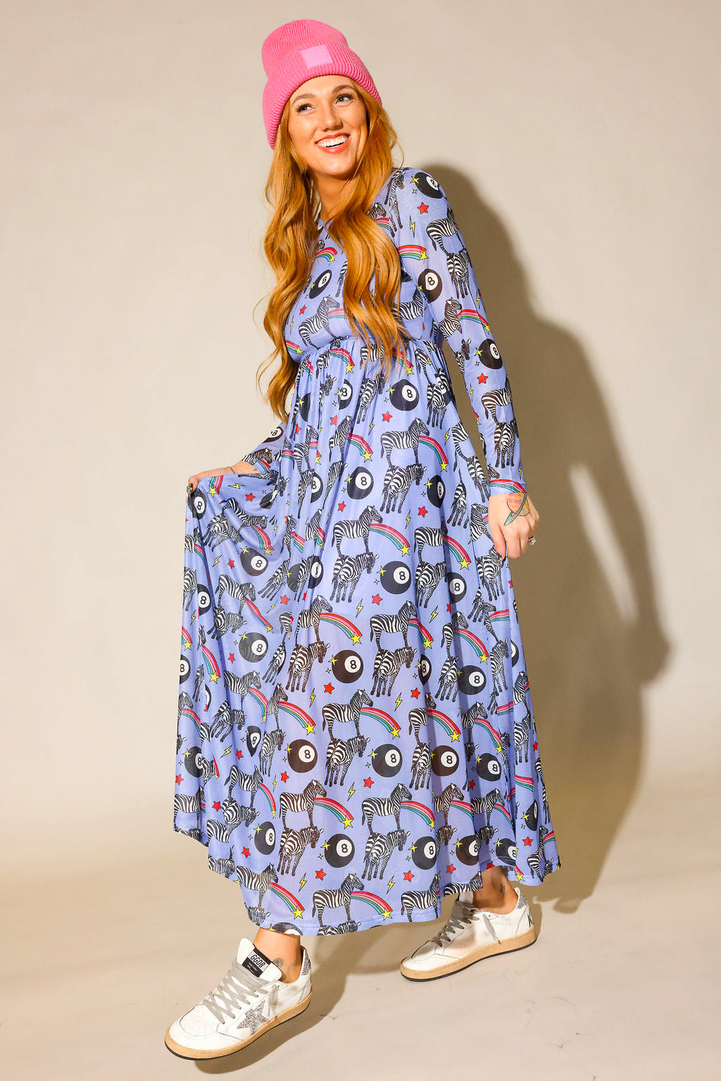 Model wearing a pink beanie, long purple mesh dress with a zebra, rainbow and 8 ball design print, and white sneakers with a silver star, against a beige background, full frontal view