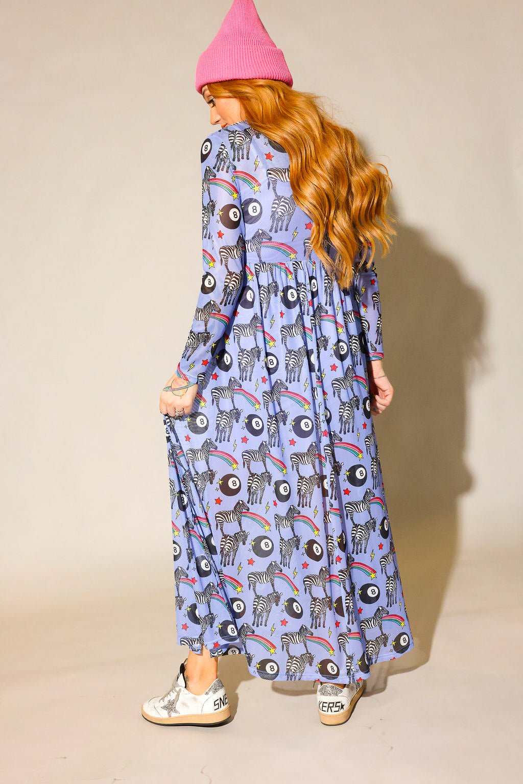 Model wearing a pink beanie, long purple mesh dress with a zebra, rainbow and 8 ball design print, and white sneakers with a silver star, on a beige background, back view