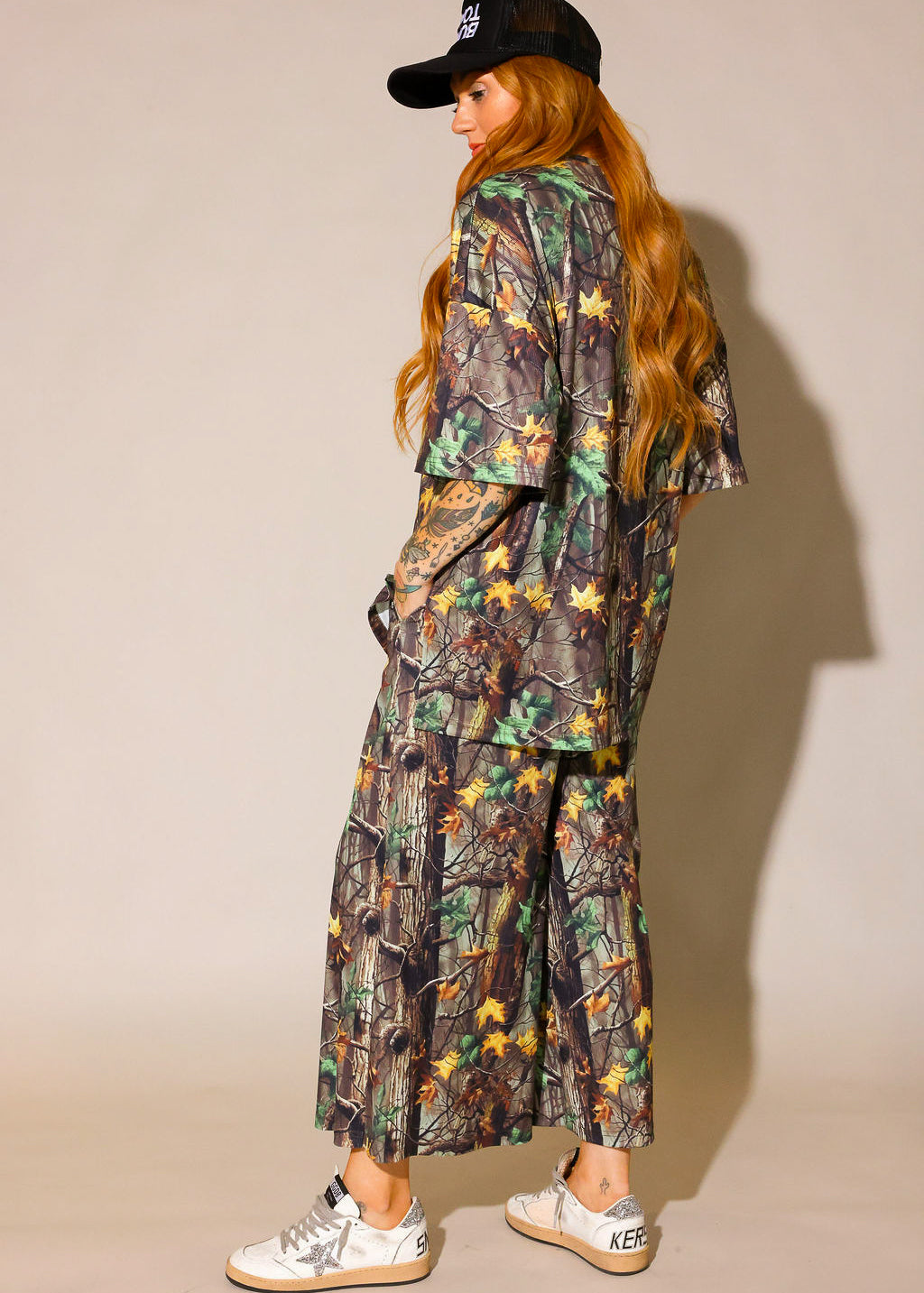 Model wearing a camouflage ribbed short sleeve and pant set with black trucker hat on a beige background, side view