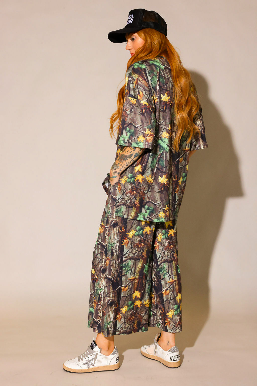 Model wearing a camouflage ribbed short sleeve and pant set with black trucker hat on a beige background, side view