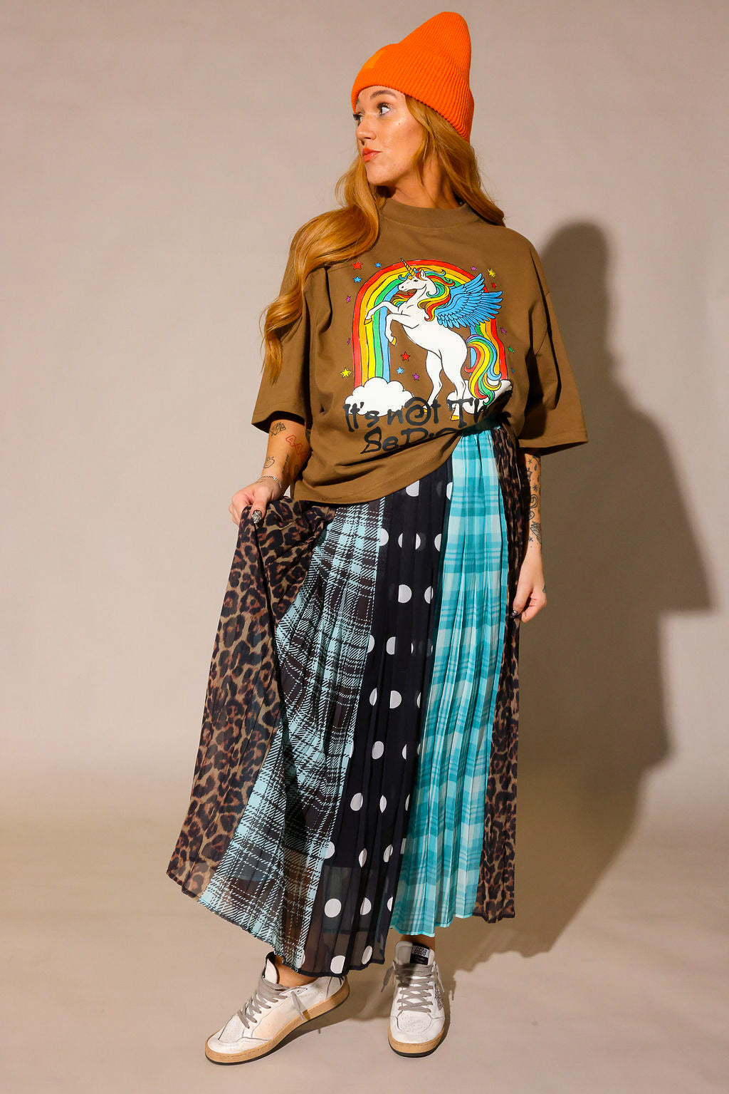 Model looking to the side, wearing a brown graphic shirt with a colorful graphic design of a rainbow and unicorn, an orange beanie, and a long, patterned skirt, with sections of black and white polka dots, blue flannel, brown flannel, and leopard prints, against a plain background, frontal view