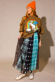 Model looking to the side, wearing a brown graphic shirt with a colorful graphic design of a rainbow and unicorn, an orange beanie, and a long, patterned skirt, with sections of black and white polka dots, blue flannel, brown flannel, and leopard prints, against a plain background, frontal view