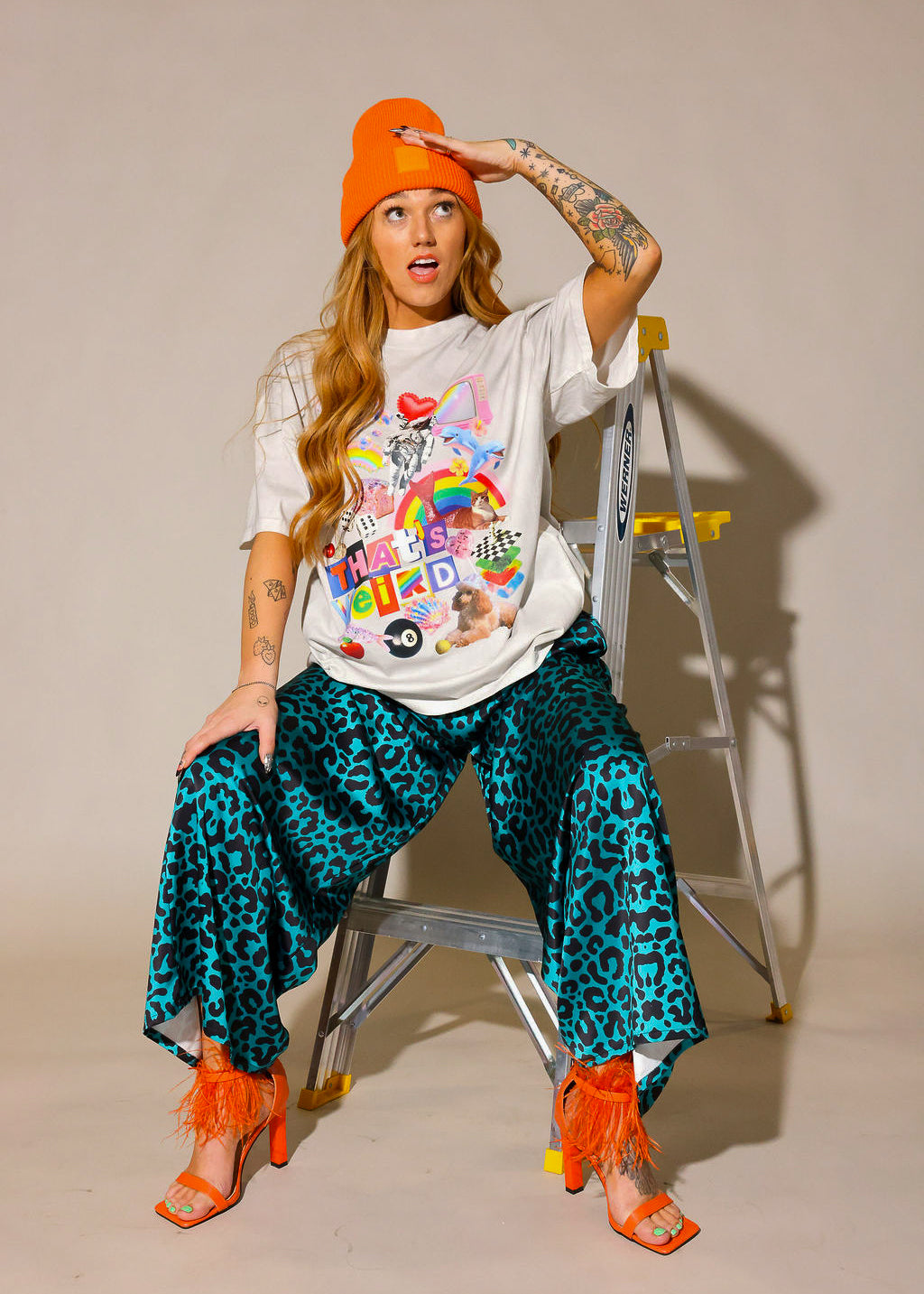 Model wearing a colorful graphic tee with a cream short sleeve shirt, leopard blue and black patterned pants, and orange high heel shoes and beanie, sitting on a ladder, frontal view