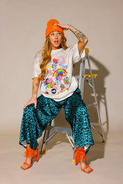 Model wearing a colorful graphic tee with a cream short sleeve shirt, leopard blue and black patterned pants, and orange high heel shoes and beanie, sitting on a ladder, frontal view