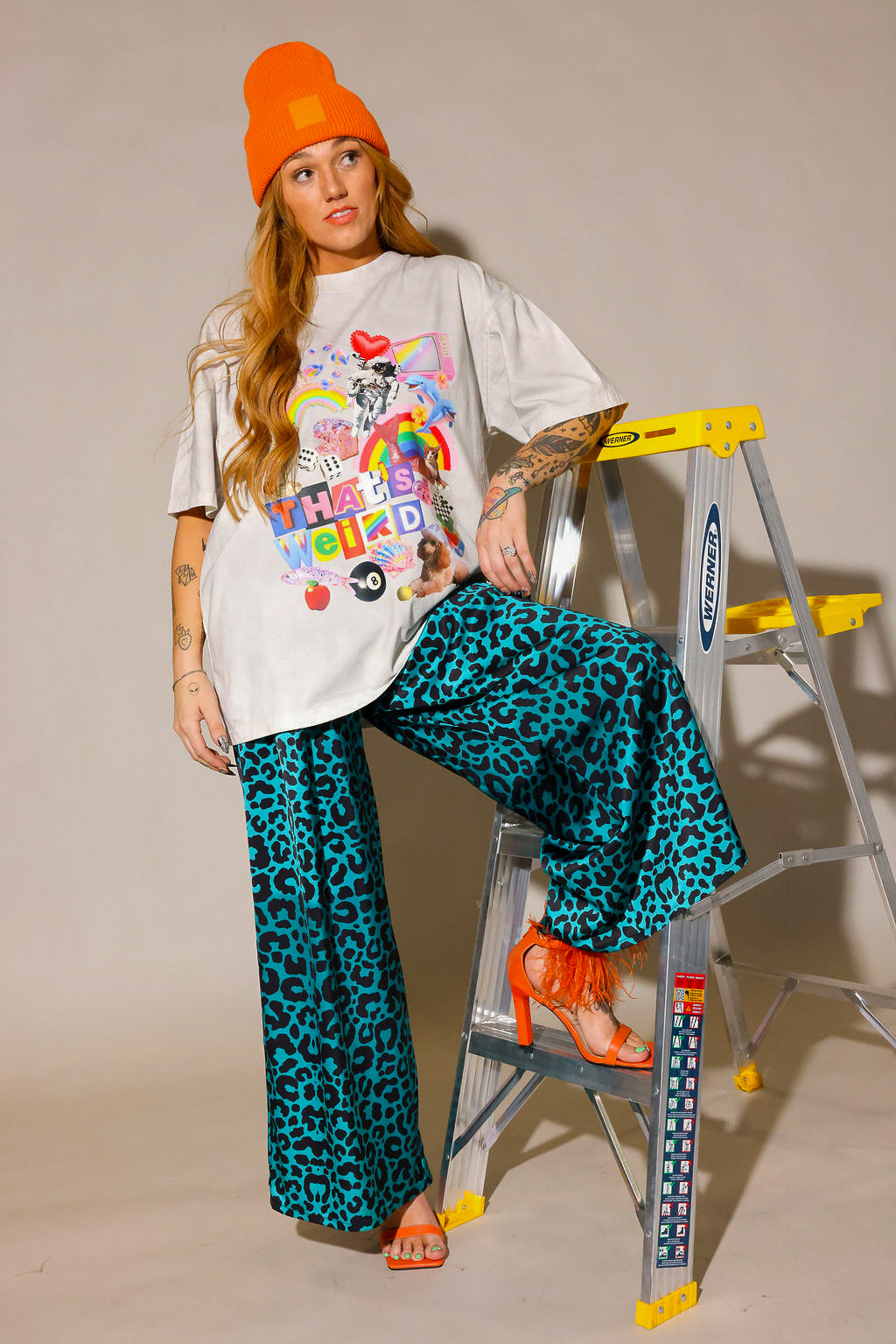Person wearing a colorful graphic tee with a cream t-shirt, leopard blue and black patterned pants, and orange high heel shoes and beanie, one foot on a ladder, frontal view