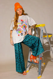Person wearing a colorful graphic tee with a cream t-shirt, leopard blue and black patterned pants, and orange high heel shoes and beanie, one foot on a ladder, frontal view