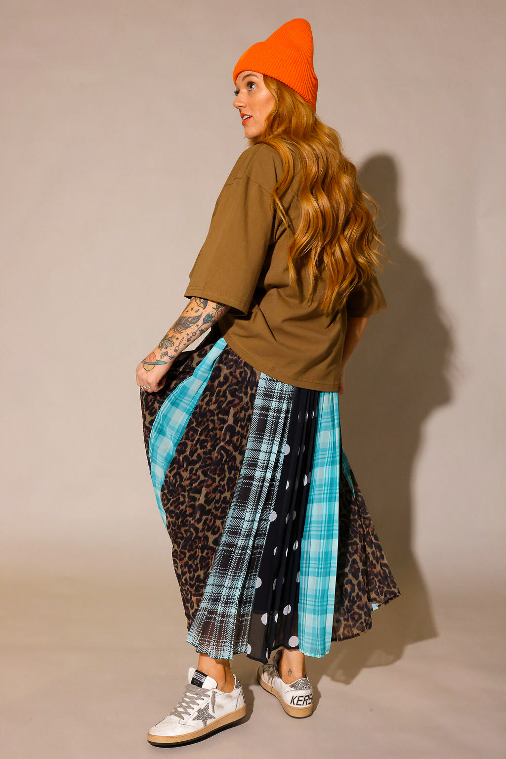 Model wearing a brown oversized tee, orange beanie and a long, patterned skirt, with sections of black and white polka dots, blue flannel, brown flannel, and leopard prints, show the back slip underneath, against a plain background, side view
