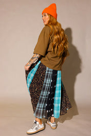 Model wearing a brown oversized tee, orange beanie and a long, patterned skirt, with sections of black and white polka dots, blue flannel, brown flannel, and leopard prints, show the back slip underneath, against a plain background, side view