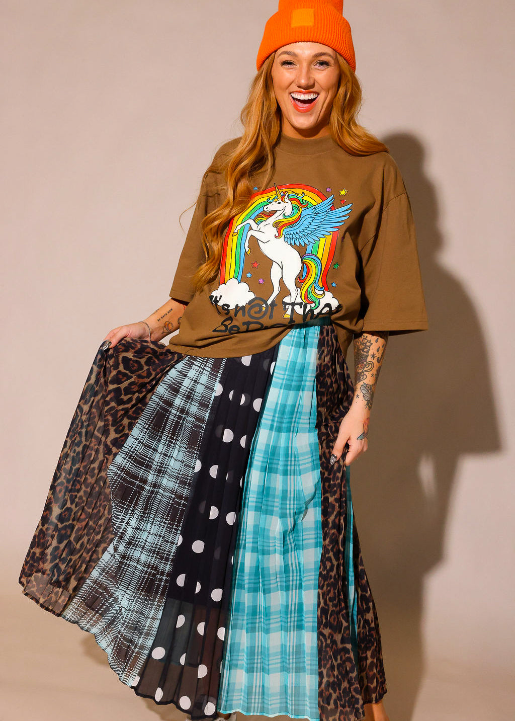 Model wearing a brown graphic shirt with a colorful graphic design of a rainbow and unicorn, an orange beanie, and a long, patterned skirt, with sections of black and white polka dots, blue flannel, brown flannel, and leopard prints, against a plain background, frontal view