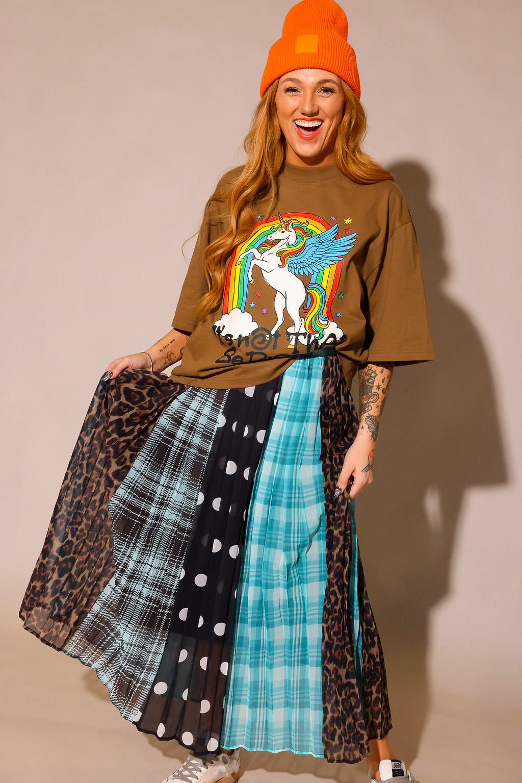 Model wearing a brown graphic shirt with a colorful graphic design of a rainbow and unicorn, an orange beanie, and a long, patterned skirt, with sections of black and white polka dots, blue flannel, brown flannel, and leopard prints, against a plain background, frontal view
